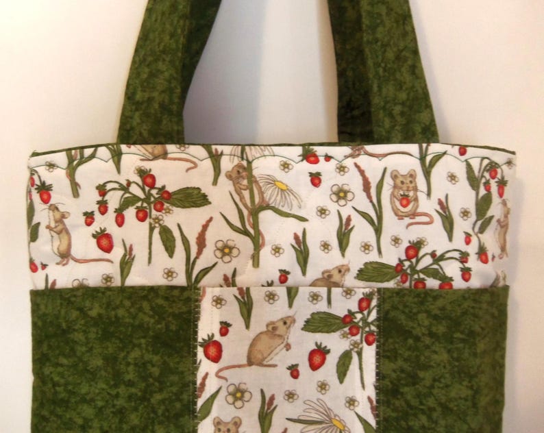 May include: A quilted tote bag featuring a white fabric panel with a repeating pattern of brown mice, red strawberries, and white flowers, contrasted by solid dark green fabric panels and matching dark green shoulder straps.
