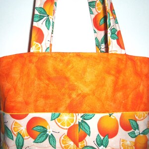 May include: A quilted tote bag with orange fabric and handles. The bag features a vibrant orange body and is accented with a patterned band of oranges, leaves, and blossoms. The handles also display the orange pattern.