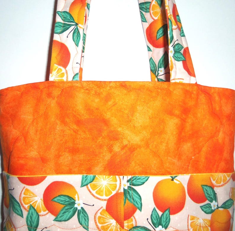 May include: A quilted tote bag with orange fabric and handles. The bag features a vibrant orange body and is accented with a patterned band of oranges, leaves, and blossoms. The handles also display the orange pattern.