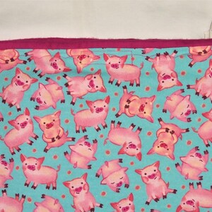 May include: A rectangular fabric item with a teal background covered in pink cartoon pigs. The pigs are in various poses. The top edge is a solid magenta color.