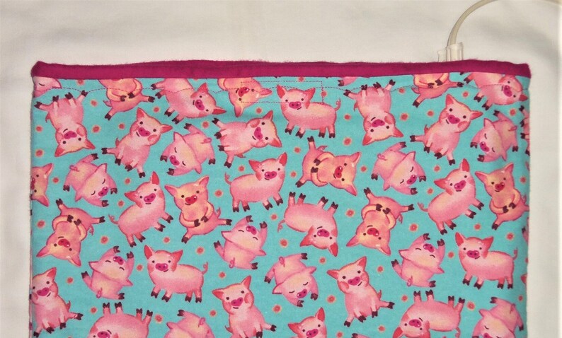 May include: A rectangular fabric item with a teal background covered in pink cartoon pigs. The pigs are in various poses. The top edge is a solid magenta color.
