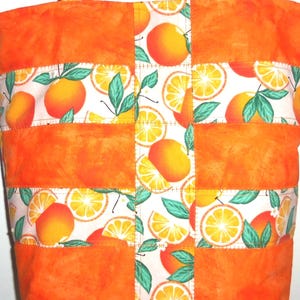 May include: Orange tote bag with a patchwork design. The bag features solid orange panels and patterned sections with orange fruit and green leaves on a white background. The bag has orange handles.