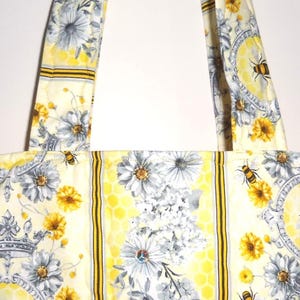 May include: A cream-colored tote bag with a floral and bee pattern. The bag features a repeating design of gray and yellow flowers, bees, and crowns. The bag has two matching straps.