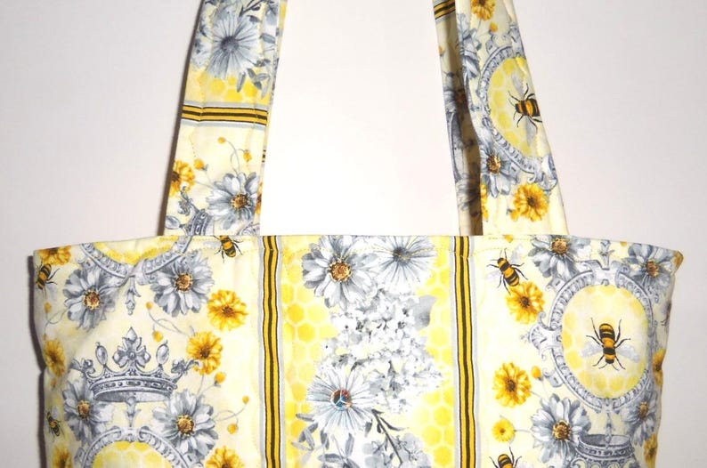 May include: A cream-colored tote bag with a floral and bee pattern. The bag features a repeating design of gray and yellow flowers, bees, and crowns. The bag has two matching straps.