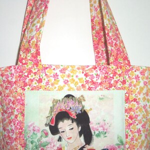 May include: A fabric tote bag featuring a vibrant floral pattern in shades of pink, orange, and yellow. The center of the bag displays a rectangular panel with an illustration of a person with dark hair adorned with flowers and hair ornaments.