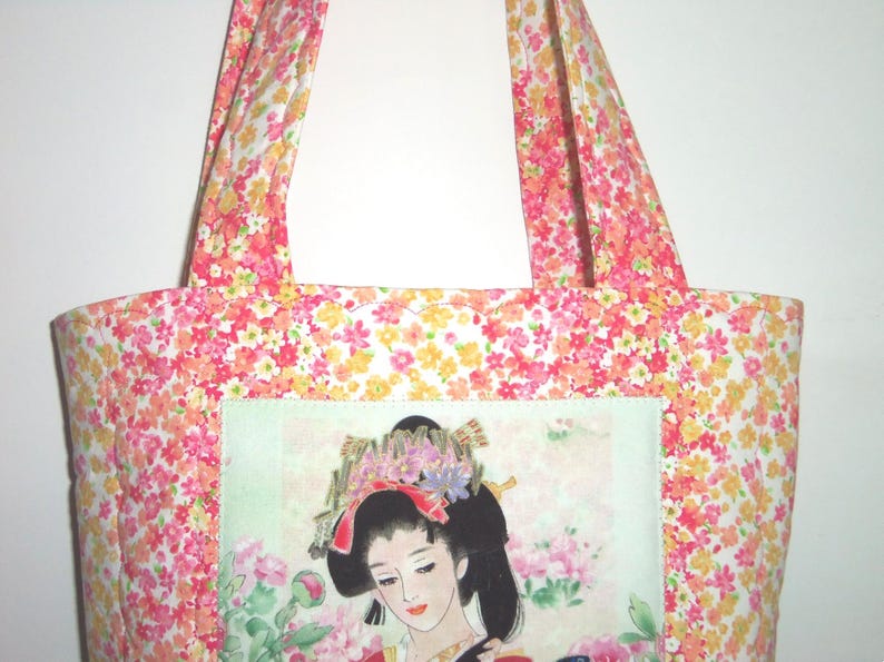 May include: A fabric tote bag featuring a vibrant floral pattern in shades of pink, orange, and yellow. The center of the bag displays a rectangular panel with an illustration of a person with dark hair adorned with flowers and hair ornaments.
