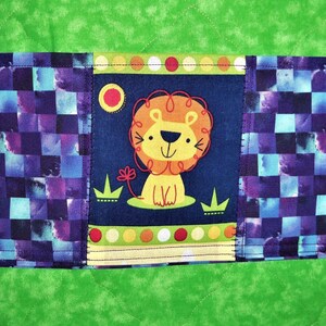May include: A quilted fabric item featuring a bright green background with a central horizontal band. This band displays a dark blue panel with a cartoon lion illustration, flanked by sections of a purple and blue checkered pattern.