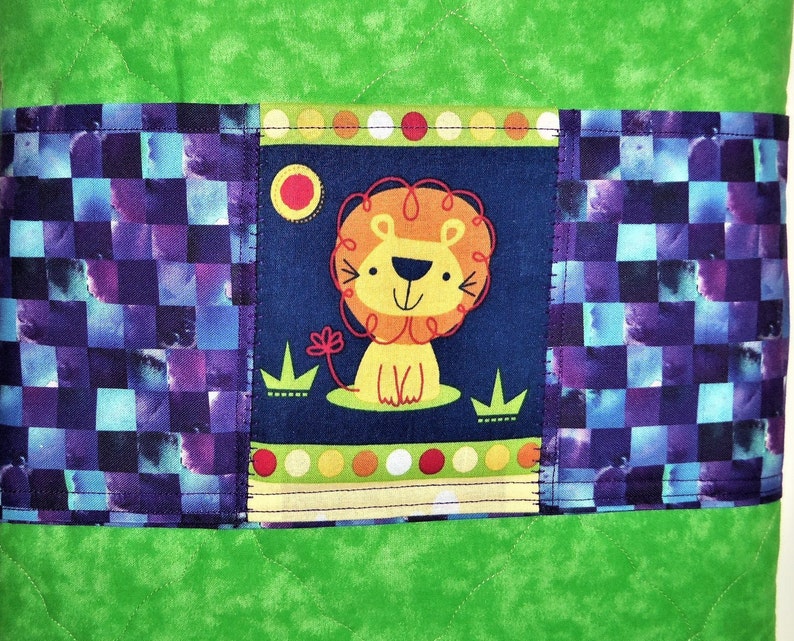 May include: A quilted fabric item featuring a bright green background with a central horizontal band. This band displays a dark blue panel with a cartoon lion illustration, flanked by sections of a purple and blue checkered pattern.
