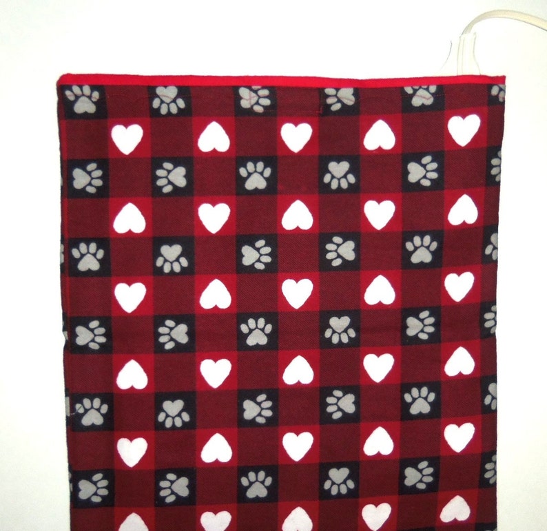 May include: Red and white checkered electric heating pad with a paw print and heart pattern. The pad is plugged in and ready to use.