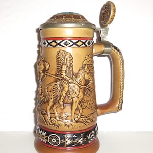 May include: A tan ceramic beer stein featuring a raised relief of a person in a feathered headdress riding a horse. The stein has a metal lid, a rope-textured handle, and decorative black, white, and red geometric bands at the top and base.