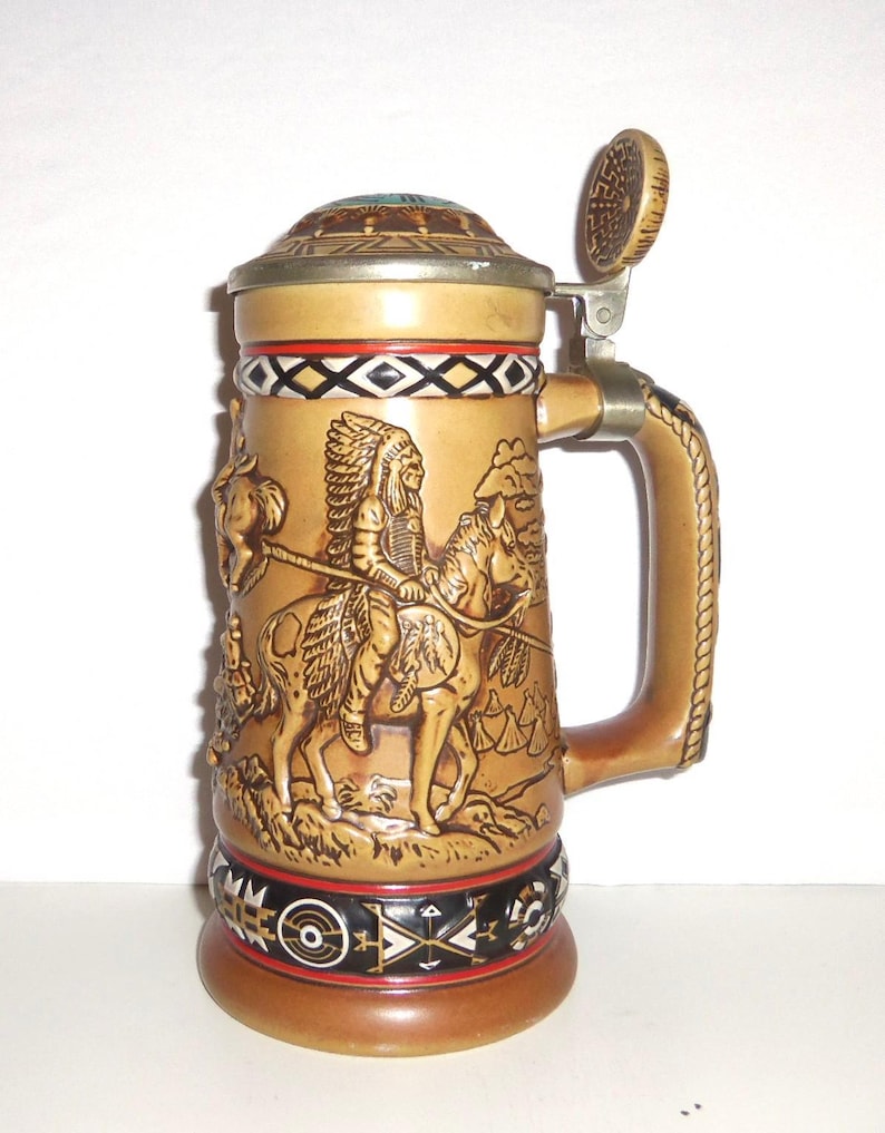 May include: A tan ceramic beer stein featuring a raised relief of a person in a feathered headdress riding a horse. The stein has a metal lid, a rope-textured handle, and decorative black, white, and red geometric bands at the top and base.