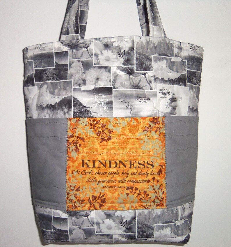 May include: A large tote bag with a patchwork design. The bag features a gray and white floral and landscape pattern, with a central orange panel that reads "KINDNESS" and a Bible verse. The bag has two handles.