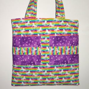 May include: A quilted tote bag featuring a vibrant rainbow striped pattern with white unicorn silhouettes. The bag includes a horizontal purple band with a subtle heart print and a vertical rainbow unicorn strip down the center with stitching.