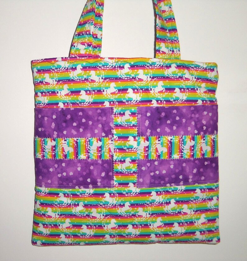 May include: A quilted tote bag featuring a vibrant rainbow striped pattern with white unicorn silhouettes. The bag includes a horizontal purple band with a subtle heart print and a vertical rainbow unicorn strip down the center with stitching.