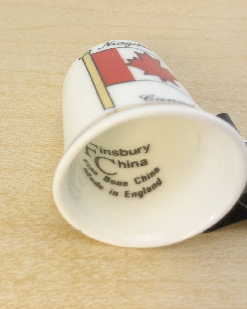 May include: White ceramic thimble with a red and white Canadian flag design. The bottom of the thimble reads "Finsbury China Fine Bone China Made in England."