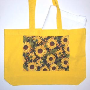 Könnte beinhalten: Eine leuchtend gelbe Tragetasche mit einer rechteckigen Vordertasche mit Sonnenblumenmuster auf dunklem Grund. Das Sonnenblumenmuster zeigt gelbe Blütenblätter und braune Mitten. Ein weißer Stoffgegenstand ist im Hauptfach sichtbar.