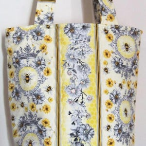 May include: A quilted tote bag with a floral and bee pattern. The bag features a cream background with yellow, gray, and black accents. The design includes flowers, bees, and decorative medallions. The handles and central stripe have a yellow honeycomb pattern.