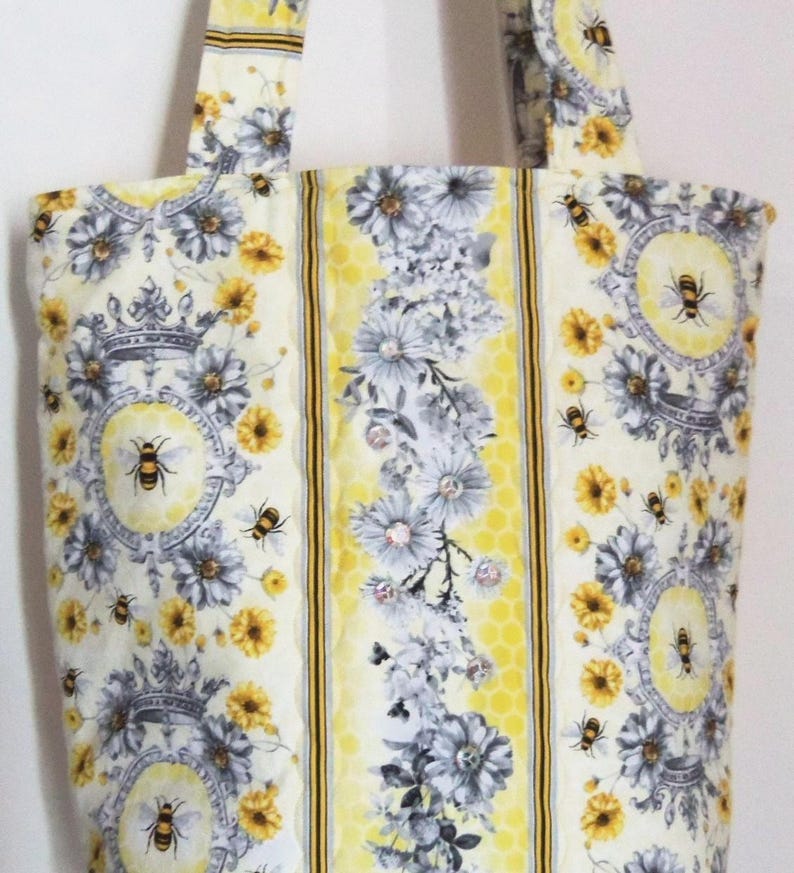 May include: A quilted tote bag with a floral and bee pattern. The bag features a cream background with yellow, gray, and black accents. The design includes flowers, bees, and decorative medallions. The handles and central stripe have a yellow honeycomb pattern.