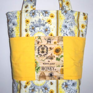 May include: A fabric tote bag with a bee and floral design. The bag features a yellow band with the text "Home is where your honey is." The bag has a floral and bee pattern in yellow, white, and gray.