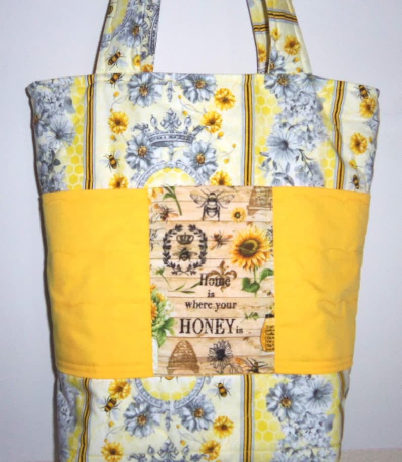 May include: A fabric tote bag with a bee and floral design. The bag features a yellow band with the text "Home is where your honey is." The bag has a floral and bee pattern in yellow, white, and gray.