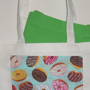May include: A white canvas tote bag featuring a rectangular front pocket with a light blue background printed with a pattern of various donuts with colorful icing and sprinkles. A bright green fabric item is tucked inside the white tote bag.