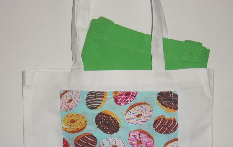 May include: A white canvas tote bag featuring a rectangular front pocket with a light blue background printed with a pattern of various donuts with colorful icing and sprinkles. A bright green fabric item is tucked inside the white tote bag.