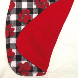 May include: A black and white buffalo plaid fabric with red paw prints. The fabric is folded over to show a red fleece lining.
