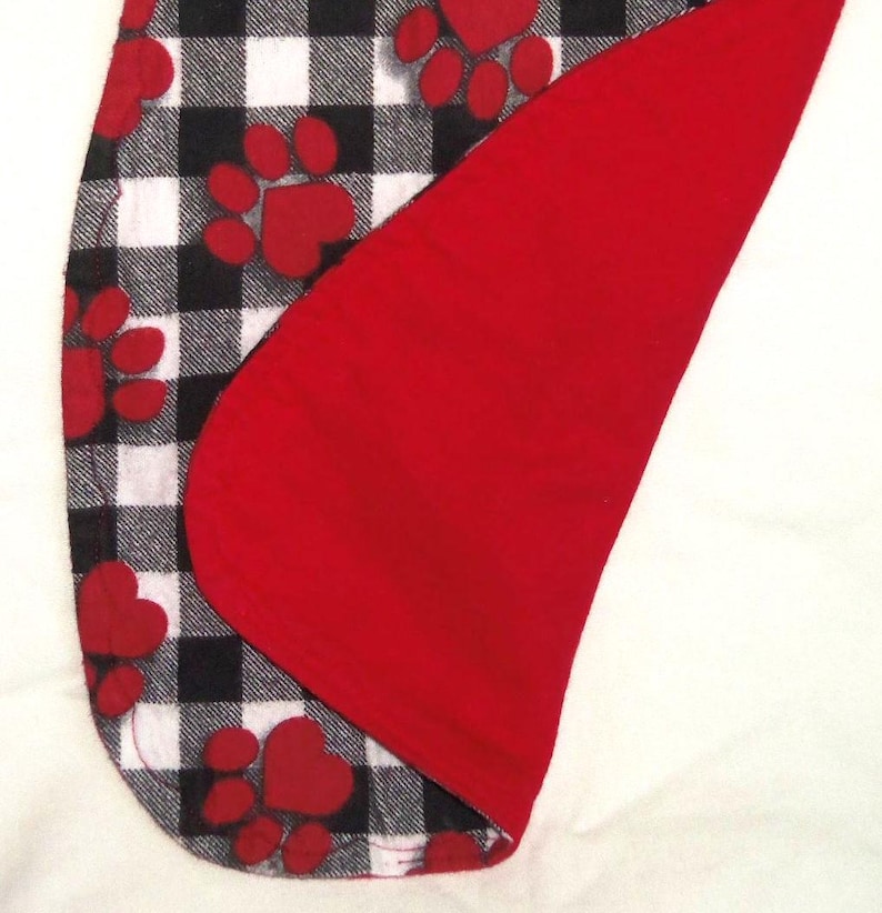 May include: A black and white buffalo plaid fabric with red paw prints. The fabric is folded over to show a red fleece lining.
