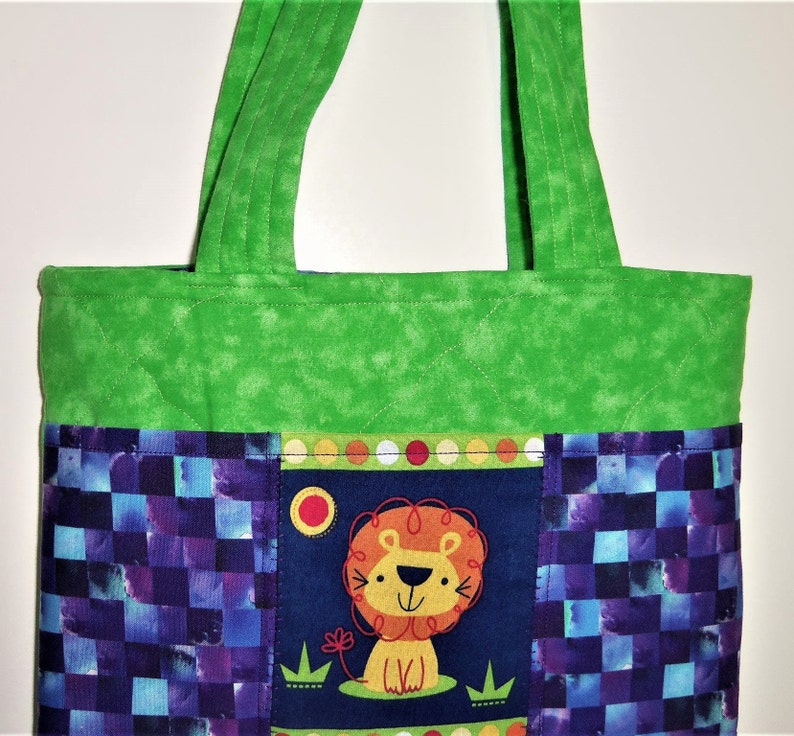 May include: A quilted tote bag featuring a bright green top section and handles. The lower portion displays a dark blue panel with a central graphic of a cartoon lion, surrounded by side panels with a purple and blue checkered pattern.
