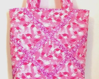 May include: A fabric tote bag featuring a repeating pattern of pink flamingos on a light pink background. The bag is accented with two diagonal strips of decorative pink floral trim that cross in the center. It includes two matching fabric handles.
