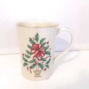 May include: A white ceramic mug featuring a gold-trimmed rim and a handle. The front displays a graphic of a green holly plant with red berries arranged in a vase, accented by a prominent red ribbon tied in a bow at the center of the foliage.