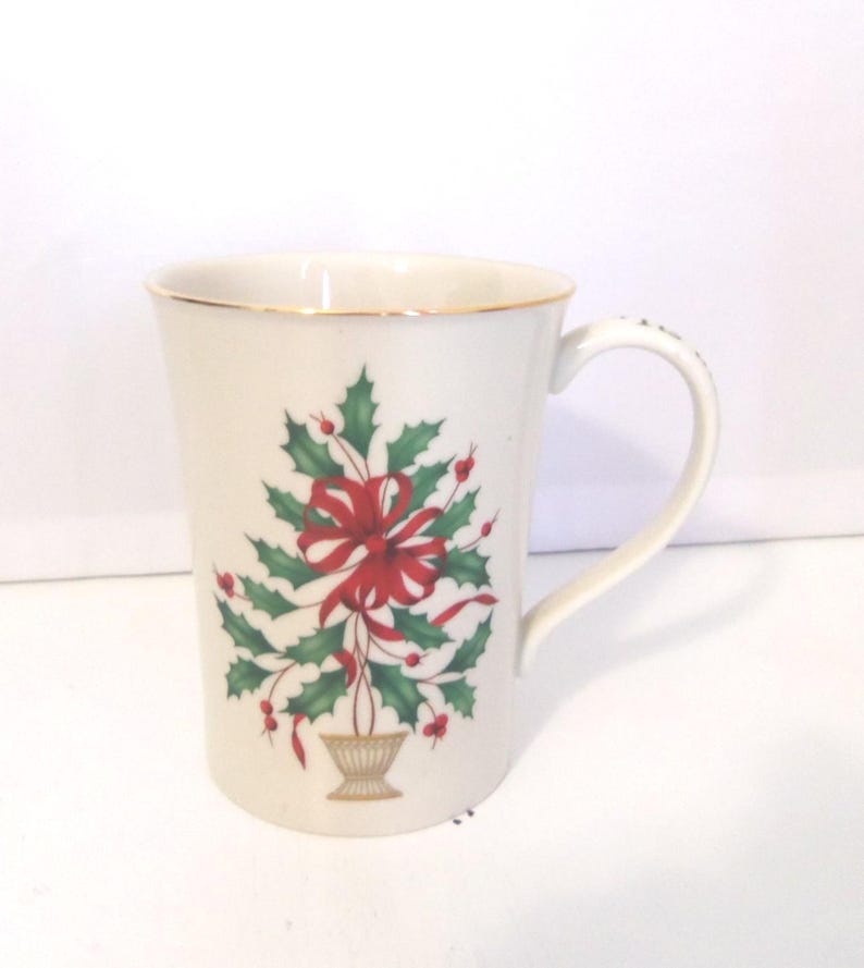 May include: A white ceramic mug featuring a gold-trimmed rim and a handle. The front displays a graphic of a green holly plant with red berries arranged in a vase, accented by a prominent red ribbon tied in a bow at the center of the foliage.