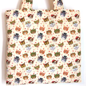 K&ouml;nnte beinhalten: Eine cremefarbene Stofftragetasche mit einem sich wiederholenden Muster aus Katzengesichtern. Die Katzen tragen verschiedene Accessoires wie Zylinder, Baskenm&uuml;tzen, Blumenkr&auml;nze und Brillen, durchsetzt mit kleinen roten Herzen und Pfotenabdr&uuml;cken.