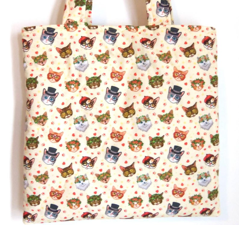 K&ouml;nnte beinhalten: Eine cremefarbene Stofftragetasche mit einem sich wiederholenden Muster aus Katzengesichtern. Die Katzen tragen verschiedene Accessoires wie Zylinder, Baskenm&uuml;tzen, Blumenkr&auml;nze und Brillen, durchsetzt mit kleinen roten Herzen und Pfotenabdr&uuml;cken.