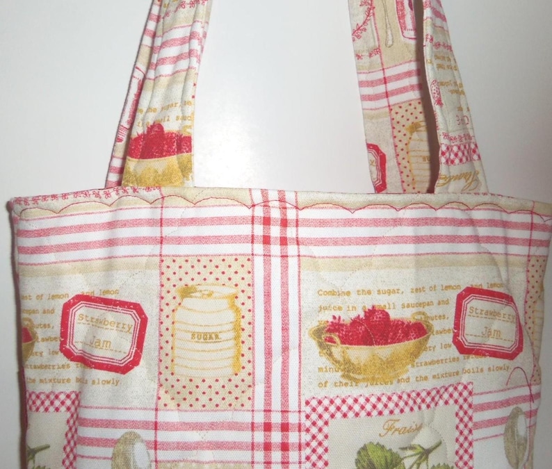 May include: A quilted fabric tote bag featuring a cream and red pattern with illustrations of strawberries, sugar canisters, and jam labels. The design includes text snippets about cooking jam and red striped sections with two matching fabric handles.
