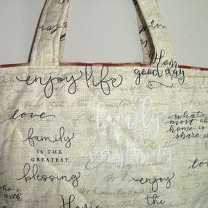May include: A beige tote bag with red trim and black script writing. The writing says "enjoy life good day", "love", "family is the greatest blessing", and "enjoy the".