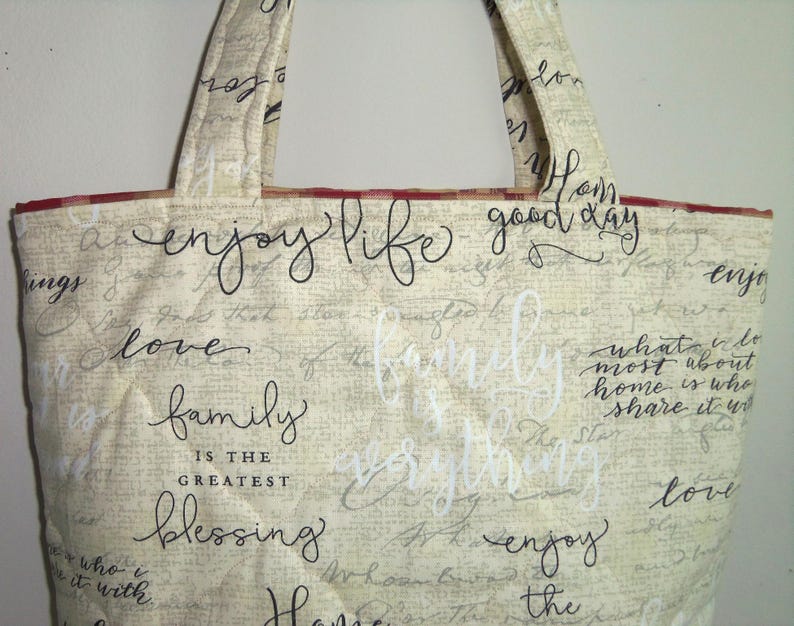 May include: A beige tote bag with red trim and black script writing. The writing says "enjoy life good day", "love", "family is the greatest blessing", and "enjoy the".