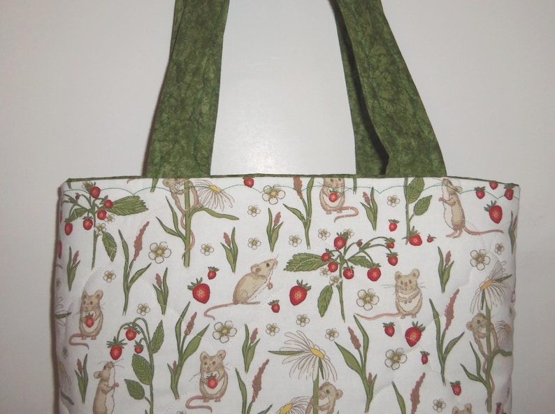 May include: A quilted tote bag featuring a white fabric base with a repeating pattern of small brown mice, red strawberries, and white flowers on green stems. The bag includes two sturdy olive green fabric handles at the top.
