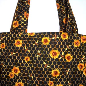 May include: A quilted tote bag featuring a black fabric base with a repeating pattern of yellow sunflowers and small bees arranged over a golden yellow honeycomb grid. The bag includes two matching fabric shoulder straps and quilted stitching.