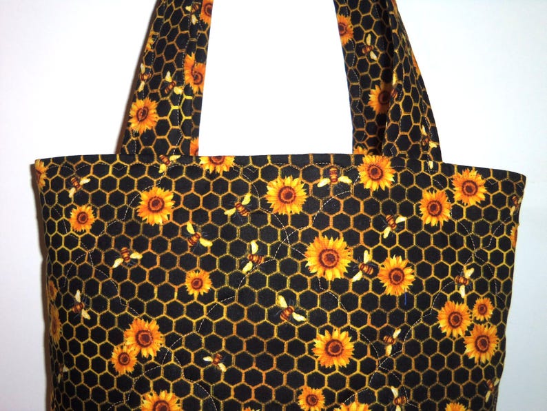 May include: A quilted tote bag featuring a black fabric base with a repeating pattern of yellow sunflowers and small bees arranged over a golden yellow honeycomb grid. The bag includes two matching fabric shoulder straps and quilted stitching.