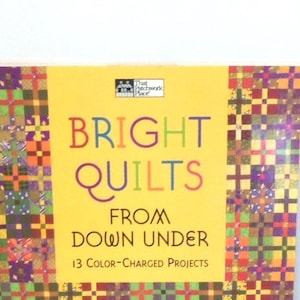 May include: A book titled Bright Quilts From Down Under with 13 Color-Charged Projects on the cover. The cover features a yellow center panel with multicolored text and a border of small, vibrant, multi-colored patchwork quilt squares.