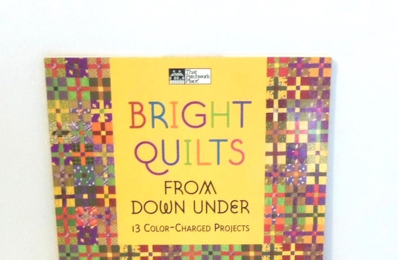 May include: A book titled Bright Quilts From Down Under with 13 Color-Charged Projects on the cover. The cover features a yellow center panel with multicolored text and a border of small, vibrant, multi-colored patchwork quilt squares.