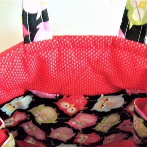 May include: A fabric tote bag featuring a bright red interior lining with a white polka dot pattern. The exterior upper section and handles display a black fabric printed with a colourful owl motif in shades of pink, green, and white.