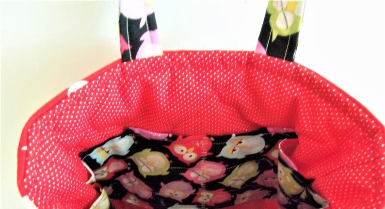 May include: A fabric tote bag featuring a bright red interior lining with a white polka dot pattern. The exterior upper section and handles display a black fabric printed with a colourful owl motif in shades of pink, green, and white.