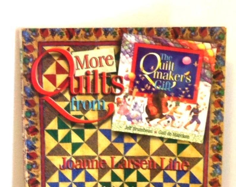K&ouml;nnte beinhalten: Ein Buch mit dem Titel More Quilts von Joanne Larsen Line mit einem Steppmuster-Cover aus gelben, gr&uuml;nen und blauen Dreiecken. Ein kleineres Buch mit dem Titel The Quiltmaker's Gift liegt oben auf dem Cover.