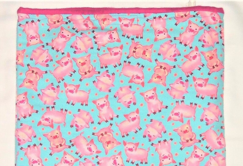 May include: A rectangular fabric with a repeating pattern of pink pigs on a light blue background with small orange dots. The edge is trimmed with a bright pink border. The pigs are in various poses.