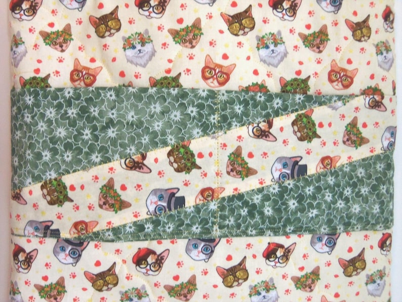 K&ouml;nnte beinhalten: Ein gesteppter Stoffbezug mit einem sich wiederholenden Muster aus Katzen, die verschiedene Accessoires wie Brillen, H&uuml;te und Blumenkr&auml;nze tragen, auf cremefarbenem Hintergrund, durchzogen von zwei diagonalen B&auml;ndern aus dunkelgr&uuml;nem Stoff mit wei&szlig;em Blumenmuster.