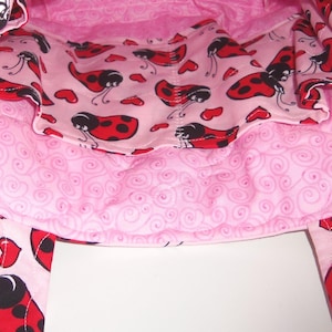 May include: A quilted fabric bag featuring a light pink interior lining with a repeating pattern of red ladybugs and small red hearts. The exterior fabric displays a light pink background with a swirling pattern and ladybug accents on the straps.