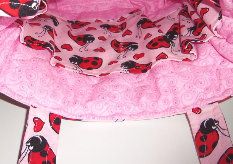 May include: A quilted fabric bag featuring a light pink interior lining with a repeating pattern of red ladybugs and small red hearts. The exterior fabric displays a light pink background with a swirling pattern and ladybug accents on the straps.