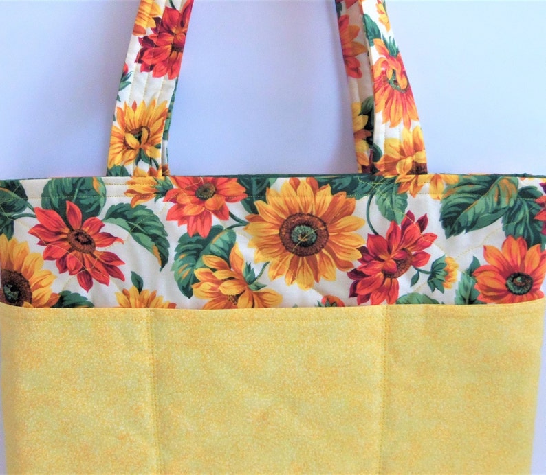 May include: A yellow tote bag with a sunflower and red flower print. The bag has a large pocket on the front.